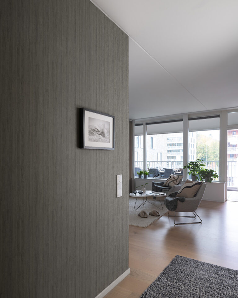 Dark wallpaper with a wood-grain pattern in a hallway adjacent to a living room with armchairs and large windows