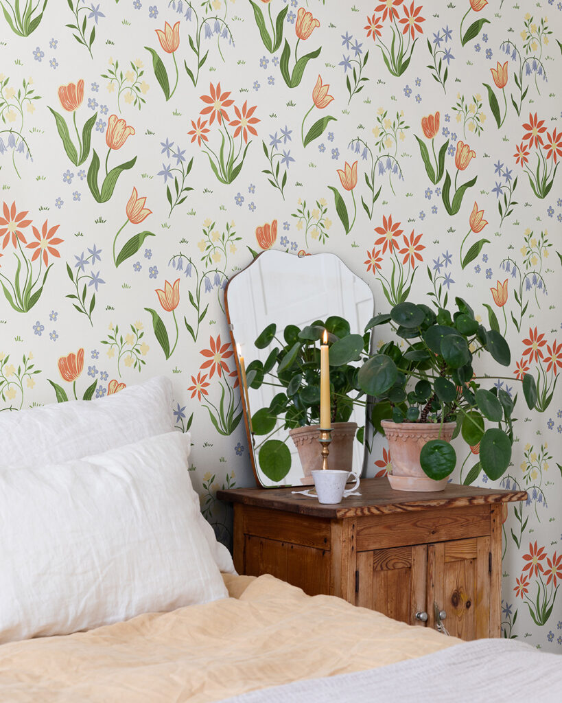 White wallpaper with colorful flowers in a bedroom, behind a bed and a potted plant