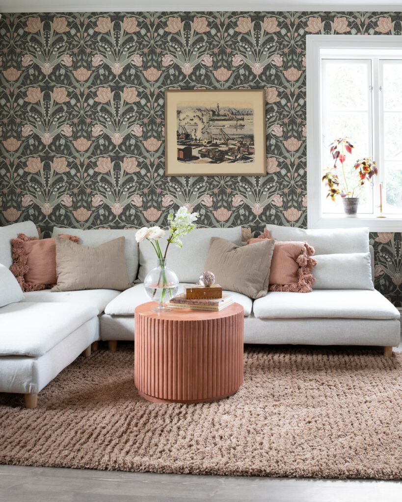 Vardagsrum med ljus soffa och gammelrosa kuddar och detaljer på väggen sitter en blommig tapet i mörka bruna och rosa toner med blommönster