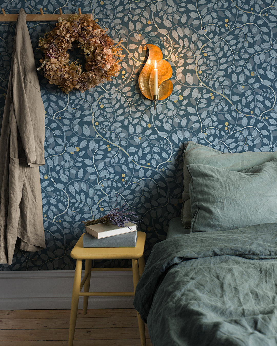 Bedroom with a made bed, a yellow stool, and a wall sconce on blue wallpaper with a leaf pattern