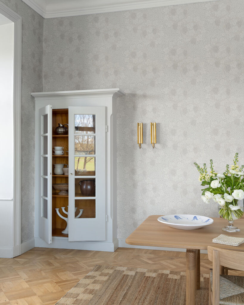 Tapet med beige botten och ljusa blad i en hall bakom ett vitt vitrinskåp