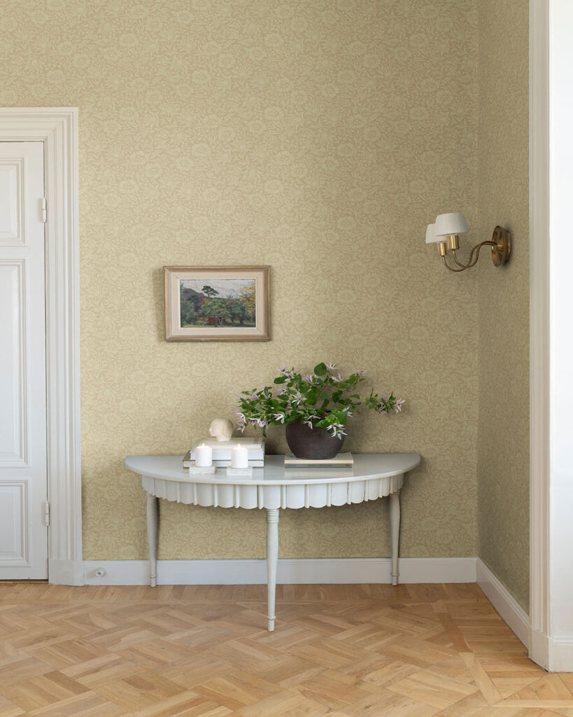 Havregul wallpaper featuring flowers and leaves in a similar color scheme in a hallway behind an old table with a plant on it